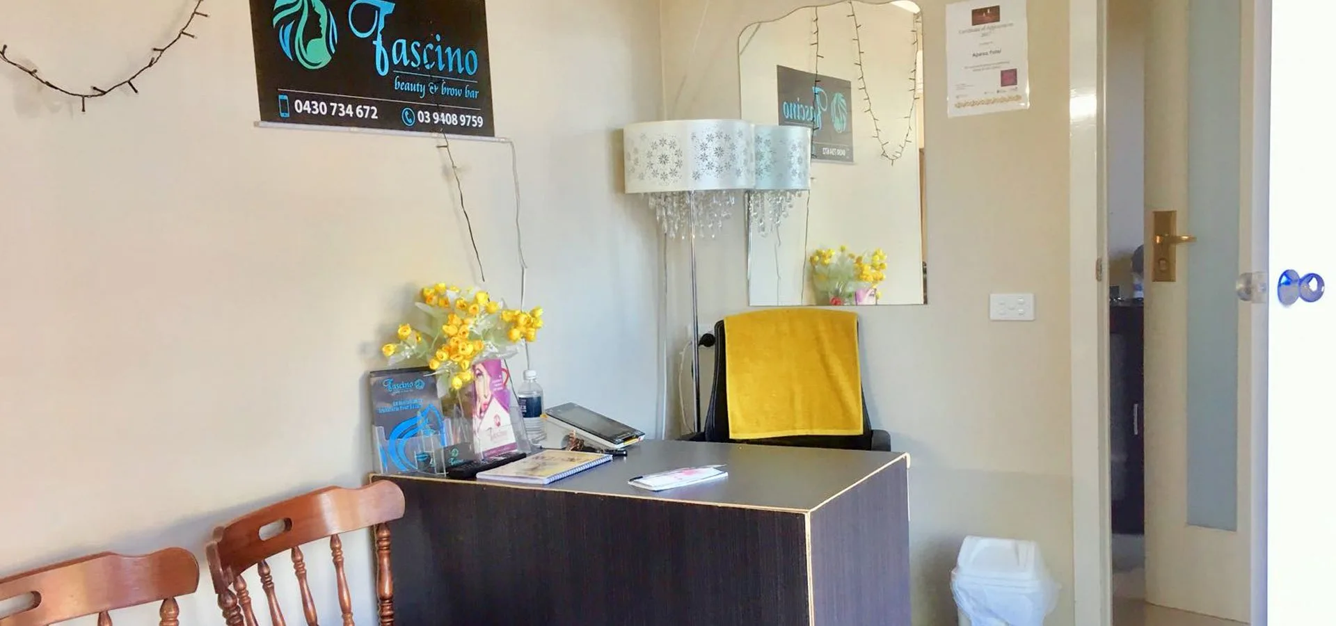 Reception corner — Fascino sign, wall mirror, black styling chair with mustard towel, floor lamp and fresh flowers